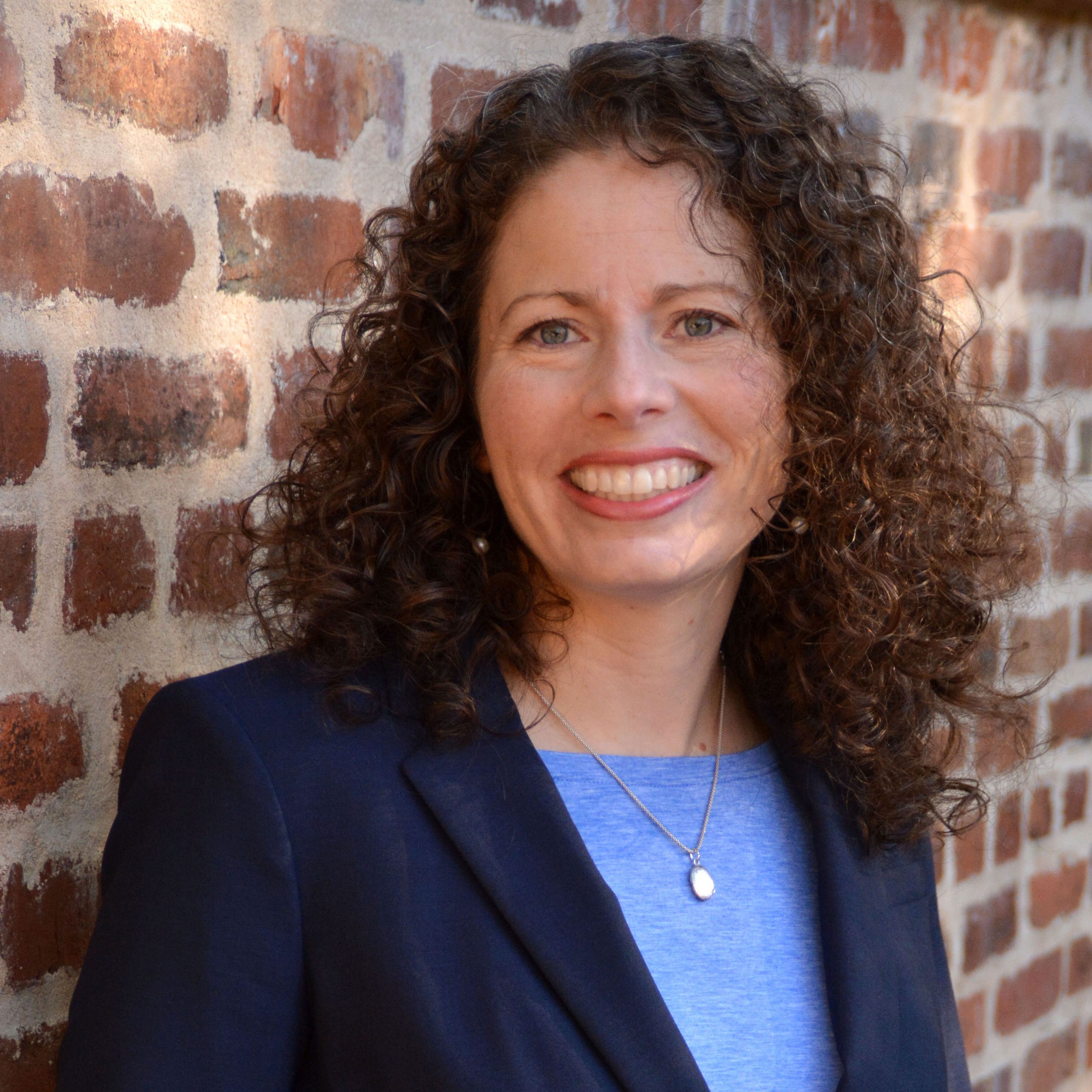A woman with curly hair smiling, wearing a dark blazer over a blue top, standing in front of a brick wall.